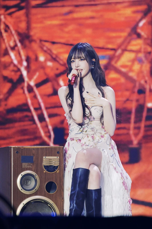 Liz singing on stage in a floral white dress with knee-high boots, seated beside a speaker.