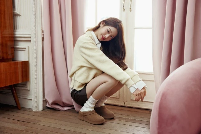 UGG campaign photograph beside pink curtains, wearing cream knitwear, a dark skirt, white socks and brown slip-on shoes.