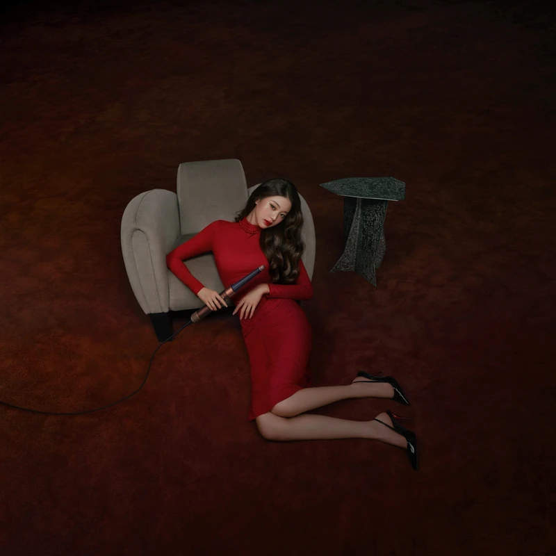 Overhead campaign photograph in a red dress beside a grey chair, holding a hair styler