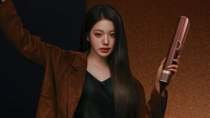A front-facing portrait in brown tailoring holding a metallic straightener